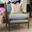 A classic English-style armchair with a dark brown wicker frame and light gray fabric cushions, styled with a patterned beige throw pillow. The chair is set on a beige rug inside a home decor showroom with floral arrangements and other furniture in the background.