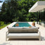 Two people sitting on a modern outdoor sofa by a pool with trees in the background